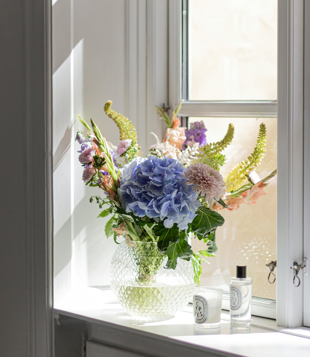 Blue hydrangeas and gladiolus in a glass vase on windowsill.